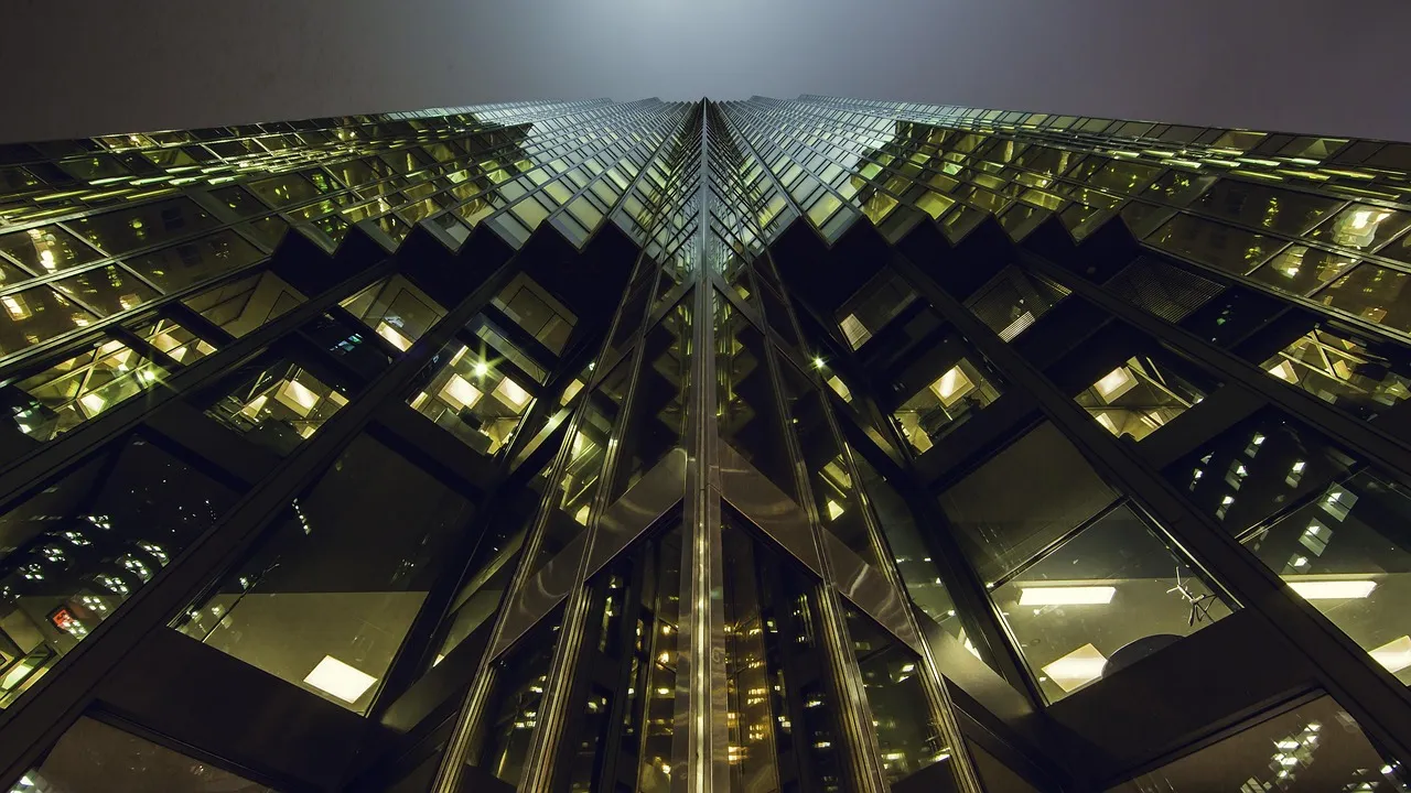 An upward, symmetrical view of a glass office tower's illuminated windows converging toward a hazy night sky.