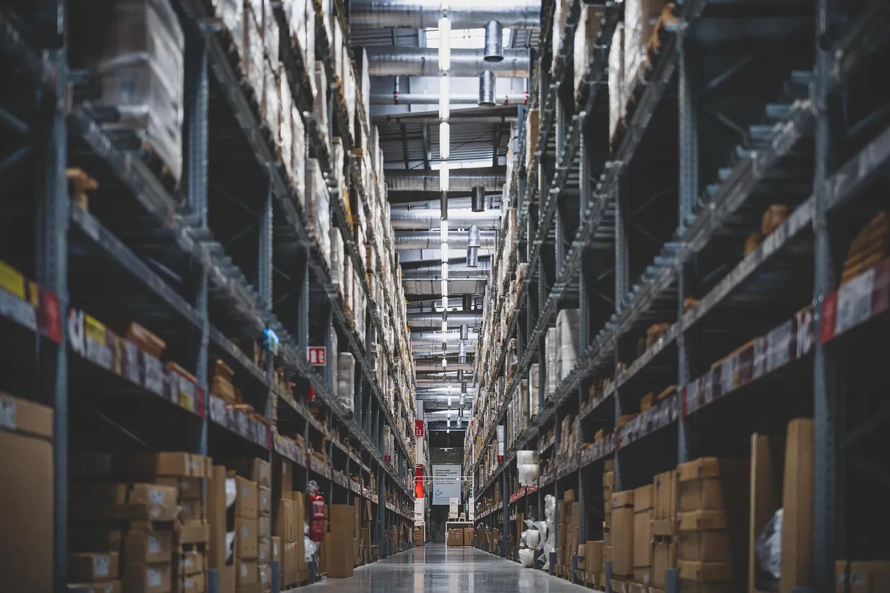 Long aisle of a warehouse retail store, tall metal racks on both sides stacked with cardboard cartons and boxed goods