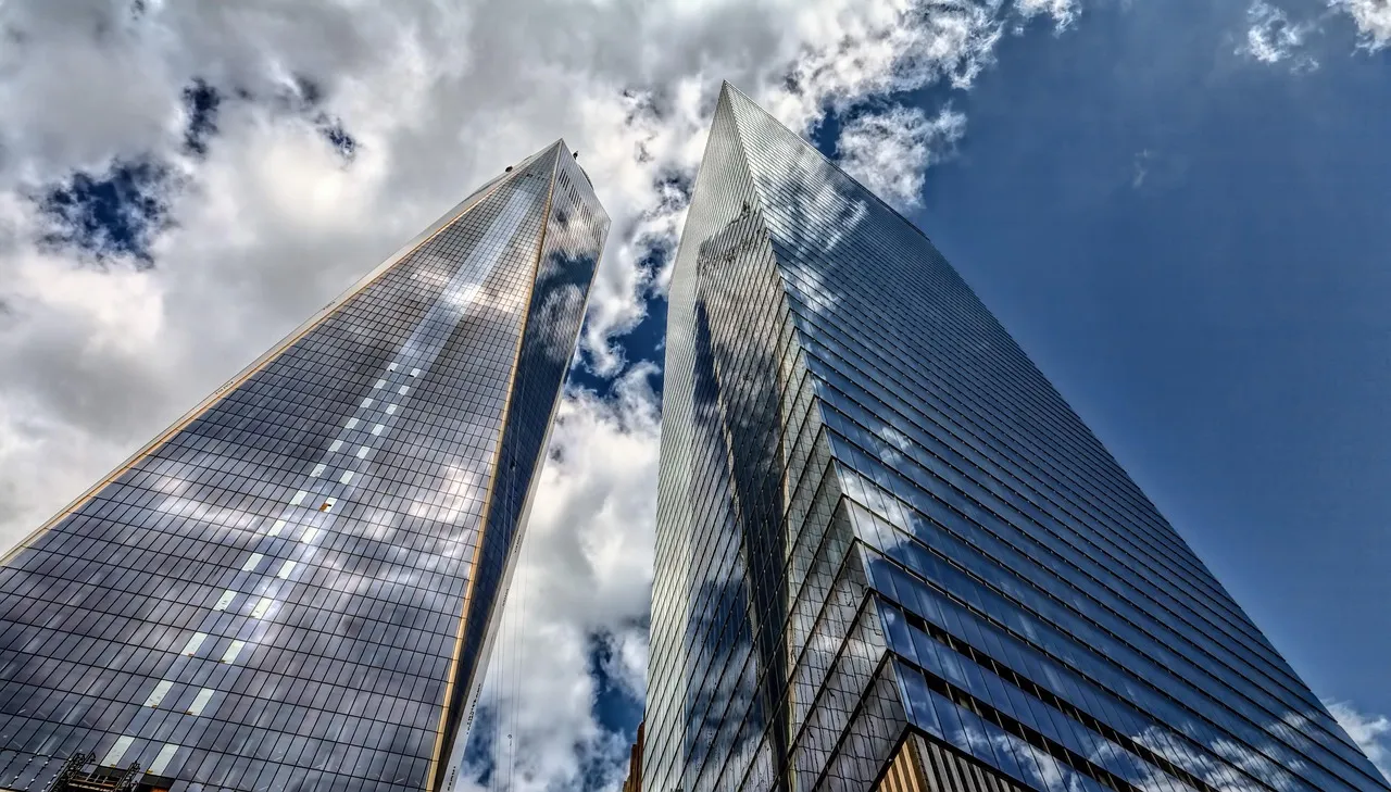 Two glass skyscrapers viewed from street level, their facades reflecting a bright sky with scattered clouds.
