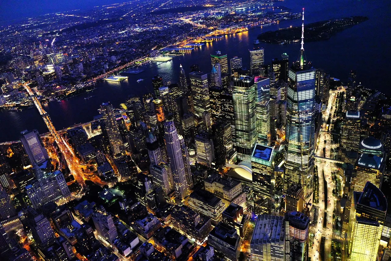 An aerial night view of Lower Manhattan, its lit skyscrapers and street grid stretching toward the rivers on either side of the island.