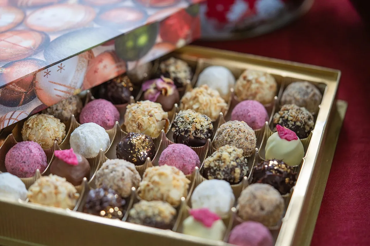 An open gold box filled with rows of assorted chocolate truffles, each in its own compartment, in varied coatings and colors