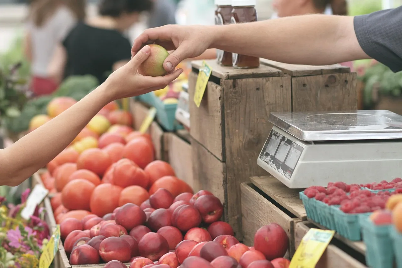 A vendor handing an apple across a farmers-market stall piled with tomatoes, nectarines, and raspberries, a scale on the counter